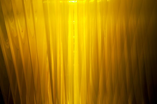 GOLD. David Spriggs. 2017. 1097 x 274 x 76 cm. / 432 x 108 x 30 inches. Yellow acrylic paint on layered sheets of transparent film, triangular gold colour structure, lighting unitsGold is the latest in David Spriggs’ chromatic artwork series of Stratachromes that examine contemporary symbolic meanings of color. Spriggs’ monumental installation presents eleven inverted yellow-golden human figures painted on layers of transparent sheets that are hung within an inverted pyramid structure. Initially reminiscent of the pediment of the New York Stock Exchange, Gold, in the spirit of pittura infamante (defaming portrait) turns the glory of capitalism on its apex, revealing its current precarious state. It speaks to the widening inequity within the Global Wealth Pyramid and the concentration of excessive wealth and corresponding power into the hands of a select few.It is fitting that this provocative art work is on display in the central business district in downtown Pittsburgh, also known as the Golden Triangle. This historic location dates back to the gilded age. It is to that age the nation’s current political leadership seek’s to return, in their quest to remake the current America into some idolized great state. But is that even possible or is such a return an illusion? And if not an illusion, will the result be to repeat that period’s excesses and failures?The viewer may be unsettled by the mirage-like-forms and suggestively paganic imagery. Is this intensely-saturated golden color representative of fools or of wealth? Yet we note that the figures of the artwork are not engaged in various forms of industry, in service to a central Goddess of Commerce, but rather are subservient, aloof or apart. Such is the relationship of many with today’s economy.